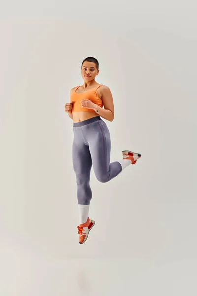 Body positivity, young short haired woman jumping on grey background, curvy fashion, female fitness, empowerment, motivation, working out, sportswear, strength and health, body image — Stock Photo