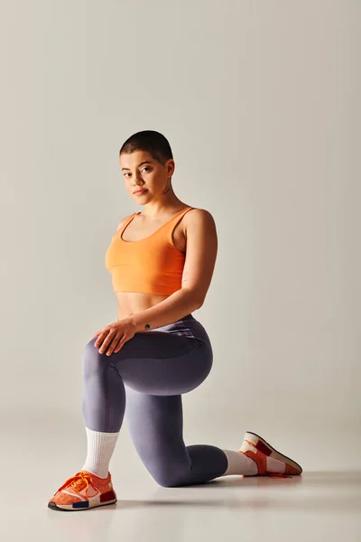Body positivity movement, young short haired woman standing on knee on grey background, curvy fitness model in sportswear, empowerment, motivation, working out with raised hands, strength and health — Stock Photo