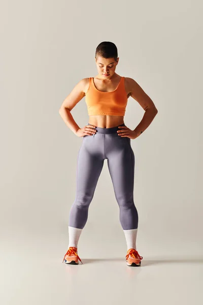 Впевненість у тілі, татуйована і короткошерста жінка, що стоїть руками на стегнах на сірому фоні, пишна фітнес-модель в спортивному одязі, розширення можливостей, мотивація, відпрацювання, сила і здоров'я — стокове фото
