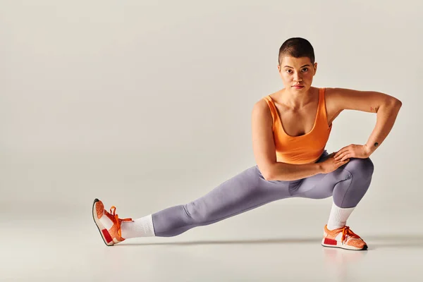 Форми тіла, гнучка і короткошерста жінка, що розтягує ноги на сірому фоні, пишна фітнес-модель в спортивному одязі, спортивний і впевнений, розширення можливостей, мотивація, відпрацювання, активний — стокове фото