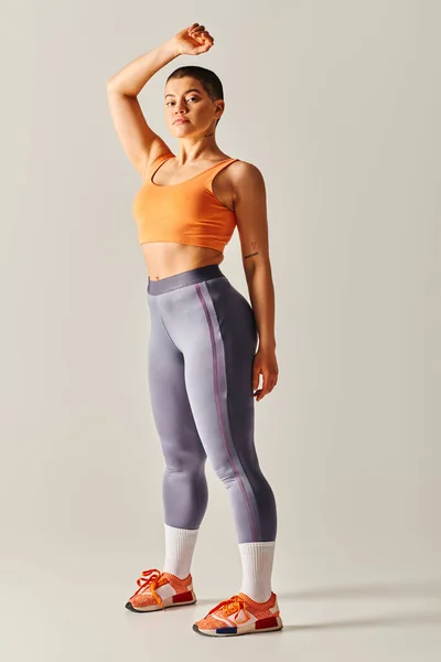 Впевненість у тілі, атлетична і короткошерста жінка позує на сірому фоні, пишна фітнес-модель, стоїть з піднятою рукою, витривалість і розширення можливостей, покоління z, повна довжина — стокове фото