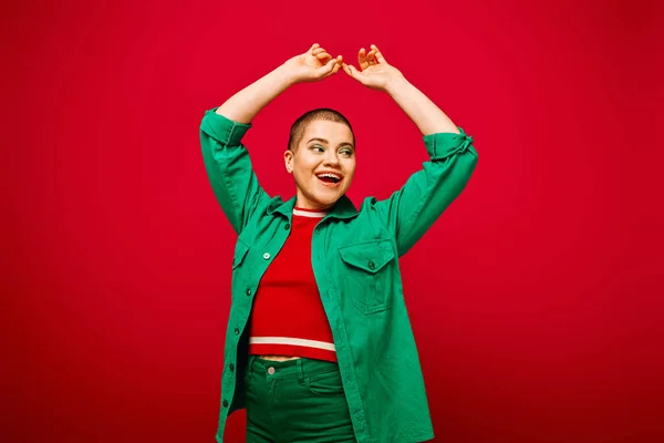 Mode und Stil, aufgeregte und kurzhaarige Frau im grünen Outfit posiert mit erhobenen Händen auf rotem Hintergrund, Generation z, Jugendkultur, moderner Hintergrund, Individualität, persönlicher Stil — Stockfoto