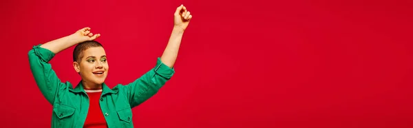 Mode und Stil, aufgeregte und kurzhaarige Frau im grünen Outfit posiert mit erhobenen Händen auf rotem Hintergrund, Generation z, Jugendkultur, moderner Hintergrund, Individualität, persönlicher Stil, Banner — Stockfoto