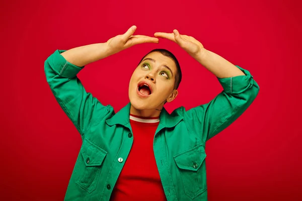 Fashion and style, amazed and short haired woman in green outfit posing with raised hands on red background, looking up generation z, youth culture, vibrant backdrop, individuality, personal style — Stock Photo