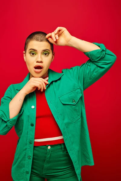 Fashion and style, shocked and short haired woman in green outfit posing with hands near face on red background, looking at camera, generation z, youth culture, vibrant backdrop, individuality — Stock Photo
