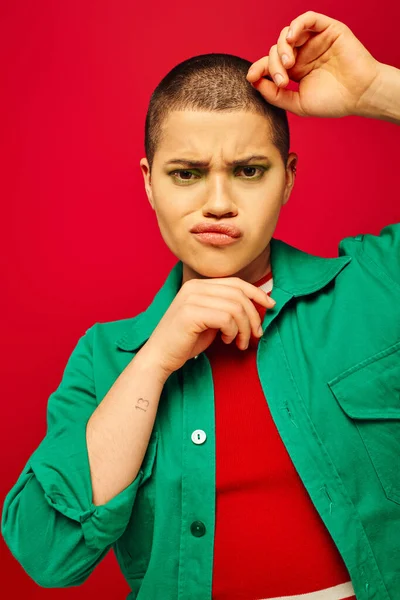 Mode und Stil, Gesichtsausdruck, unzufriedene und kurzhaarige Frau im grünen Outfit posiert auf rotem Hintergrund, blickt in die Kamera, Generation Z, Jugendkultur, lebendige Kulisse, Individualität — Stockfoto