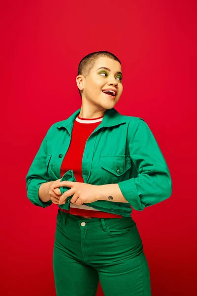 Mode und Stil, tätowierte, staunende und kurzhaarige Frau im grünen Outfit posiert auf rotem Hintergrund, schaut weg, Generation Z, Jugendkultur, lebendige Kulisse, Individualität — Stockfoto