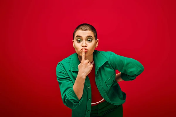 Fashion and style, tattooed, short haired woman in green outfit posing with finger near lips on red background, looking at camera, generation z, youth culture, vibrant backdrop, individuality — Stock Photo