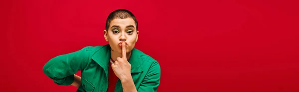 Fashion and style, tattooed, short haired woman in green outfit posing with finger near lips on red background, looking at camera, generation z, youth culture, vibrant backdrop, individuality, banner — Stock Photo
