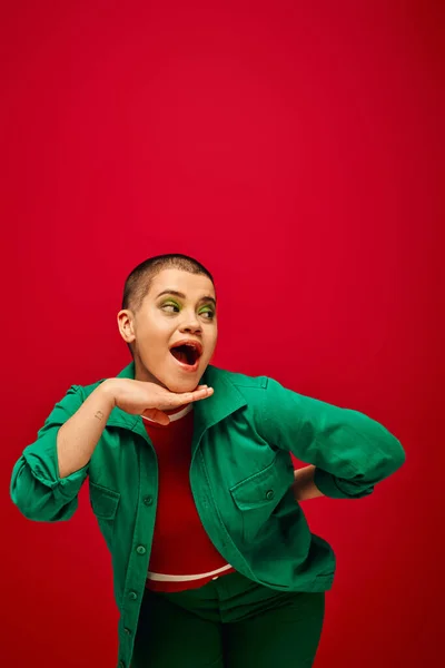 Mode und Stil, tätowiert und erstaunt, junge kurzhaarige Frau im grünen Outfit posiert auf rotem Hintergrund, schaut weg, Generation z, Jugendkultur, lebendige Kulisse, Individualität — Stockfoto