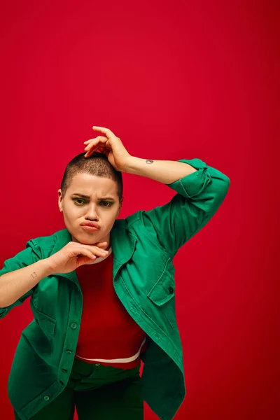 Fashion and style, emotional and tattooed, short haired woman in green outfit pouting lips on red background, looking at camera, generation z, youth culture, vibrant backdrop, individuality — Stock Photo