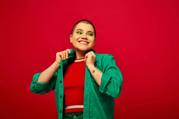 Fashion statement, happy and tattooed, short haired woman in green outfit smiling on red background, looking away, generation z, youth culture, vibrant backdrop, individuality — Stock Photo