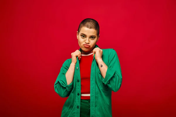 Fashion choices, emotional and tattooed, short haired woman in green outfit pouting lips on red background, looking at camera, generation z, youth culture, vibrant backdrop, personal style — Stock Photo