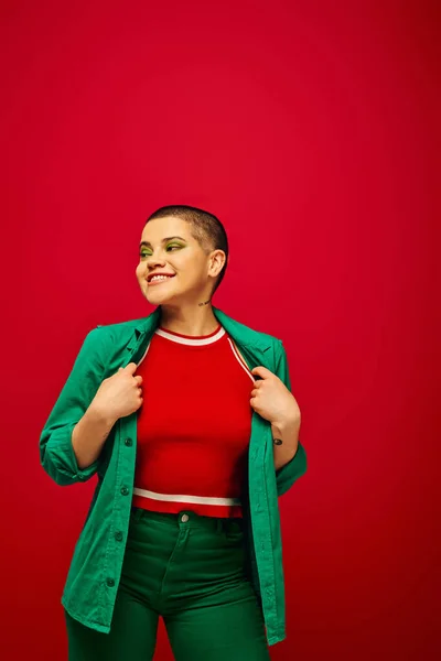 Modetrend, glücklich und tätowiert, kurzhaarige Frau im grünen Outfit, lächelnd auf rotem Hintergrund, wegschauen, Generation Z, Jugendkultur, lebendige Kulisse, Individualität, persönlicher Stil — Stockfoto