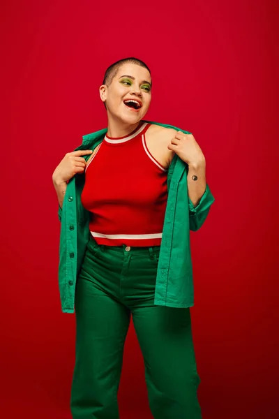 Модный тренд, возбужденная и татуированная, коротковолосая женщина в зеленом наряде, улыбающаяся на красном фоне, отводящая взгляд, поколение Z, молодость, яркий задник, индивидуальность, личный стиль — стоковое фото