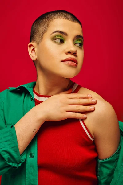 Fashion trend, young and tattooed, short haired woman in green outfit posing on red background, looking away, generation z, youth, vibrant backdrop, bold makeup, personal style, portrait — стоковое фото