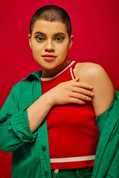 Modetrend, jugendlich und tätowiert, kurzhaarige Frau in grünem Outfit posiert auf rotem Hintergrund, blickt in die Kamera, Generation Z, Jugend, lebendige Kulisse, fettes Make-up, persönlicher Stil, Porträt — Stockfoto