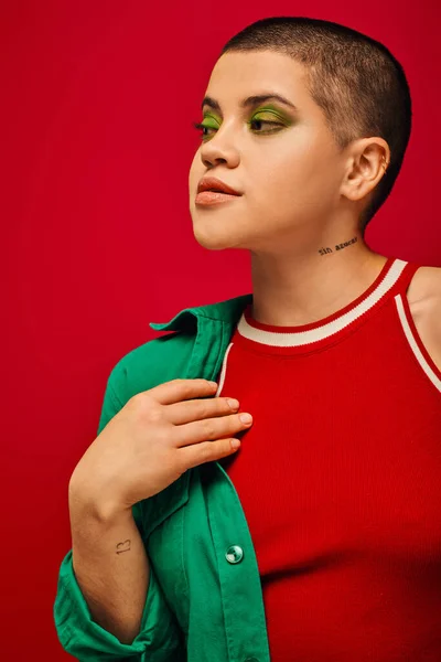 Tendencia de la moda, joven y tatuado, mujer de pelo corto en traje verde posando sobre fondo rojo, mirando hacia otro lado, generación z, juventud, telón de fondo vibrante, maquillaje en negrita, retrato - foto de stock