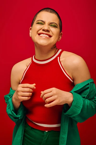 Moda tendência, alegre e tatuado, mulher de cabelos curtos em roupa verde posando no fundo vermelho, geração z, juventude, pano de fundo vibrante, maquiagem ousada, estilo pessoal, retrato — Fotografia de Stock
