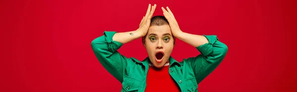 Fashion trend, shocked and tattooed, short haired woman in green outfit touching head on red background, looking away, generation z, youth, vibrant backdrop, individuality, personal style, banner — Stock Photo