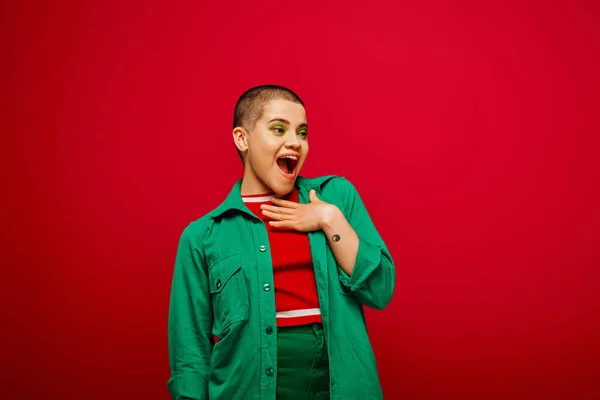 Fashion trend, amazed and tattooed, short haired woman in green outfit touching chest on red background, looking away, generation z, youth, vibrant backdrop, individuality, personal style — Stock Photo