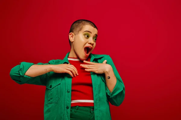 Fashion statement, amazed and tattooed, short haired woman in green outfit touching chest on red background, looking away, generation z, youth, vibrant backdrop, individuality, personal style — Stock Photo