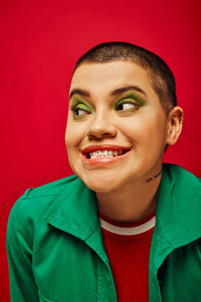 Maquillaje audaz, alegre y tatuado, mujer de pelo corto en traje verde sonriendo sobre fondo rojo, mirando hacia otro lado, generación z, juventud, telón de fondo vibrante, individualidad, estilo personal - foto de stock