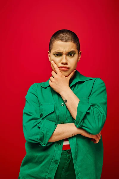 Fashion choices, displeased and tattooed, short haired woman in green outfit touching chin on red background, looking at camera, generation z, youth, vibrant backdrop, individuality, personal style — Stock Photo