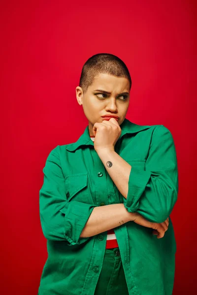 Fashion choices, displeased and tattooed, short haired woman in green outfit touching chin on red background, looking away, generation z, youth, vibrant backdrop, emotional and youthful — Stock Photo