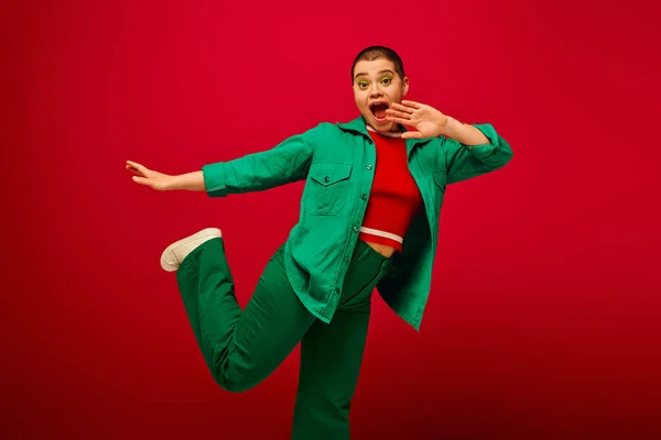 Stylish look, amazed and tattooed, short haired woman in green outfit gesturing on red background, looking at camera, generation z, youth, vibrant backdrop, individuality, personal style — Stock Photo