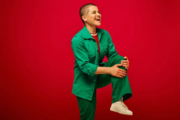 Stylish look, bold makeup, cheerful and tattooed, short haired woman in green outfit posing on red background, looking away, generation z, youth, vibrant backdrop, individuality, personal style — Stock Photo