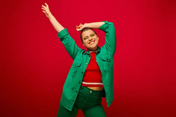 Stylisches Outfit, fettes Make-up, fröhlich und tätowiert, kurzhaarige Frau im grünen Outfit posiert auf rotem Hintergrund, Generation Z, Jugendkultur, lebendiger Hintergrund, persönlicher Stil — Stockfoto