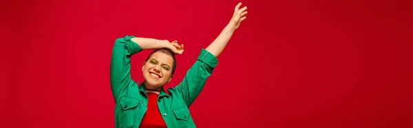 Traje elegante, maquillaje audaz, alegre y tatuado, mujer de pelo corto en traje verde posando sobre fondo rojo, generación z, cultura juvenil, telón de fondo vibrante, estilo personal, bandera - foto de stock