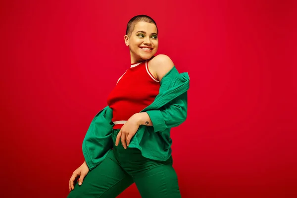 Stylish appearance, bold makeup, happy and tattooed, short haired woman in green outfit posing on red background, generation z, youth, vibrant backdrop, individuality, personal style — Stock Photo