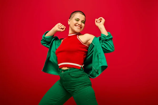 Youth culture, stylish appearance, bold makeup, happy and tattooed, short haired woman in green outfit dancing on red background, generation z, youth, vibrant backdrop, individuality, personal style — Stock Photo
