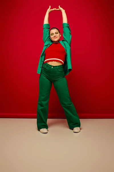 Fashion statement, vibrant backdrop, cheerful and young woman in stylish attire posing with raised hands on red background, full length, generation z, youth culture, personal style, curvy fashion — Stock Photo