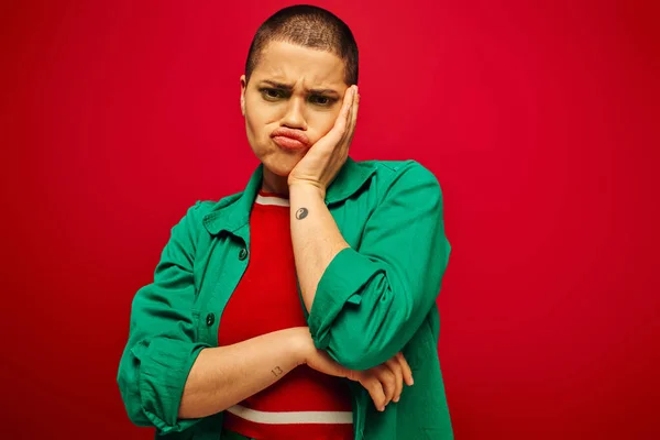 Declaración de moda, maquillaje audaz, generación z, cultura juvenil, mujer joven triste con los labios de pelo corto pucheo sobre fondo rojo, ropa casual, cultura juvenil, fondo vibrante, aspecto elegante - foto de stock