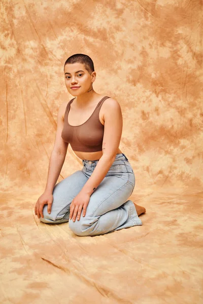 Representation of body, acceptance, curvy young and tattooed woman in jeans and crop top sitting on mottled beige background, personal style, self-acceptance, generation z, denim fashion, tattooed — Stock Photo