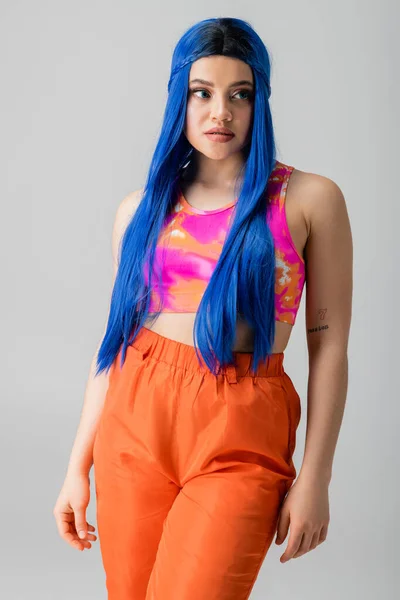 Moda hacia adelante, energía juvenil, mujer joven tatuada con el pelo azul posando en ropa de colores sobre fondo gris, individualismo, estilo moderno, moda urbana, color vibrante, modelo femenino - foto de stock