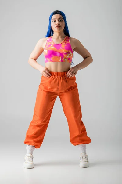 Fashion, tattooed young woman with blue hair posing with hands on hips and standing in colorful clothes on grey background, full length, individualism, modern style, urban fashion, vibrant color — Stock Photo