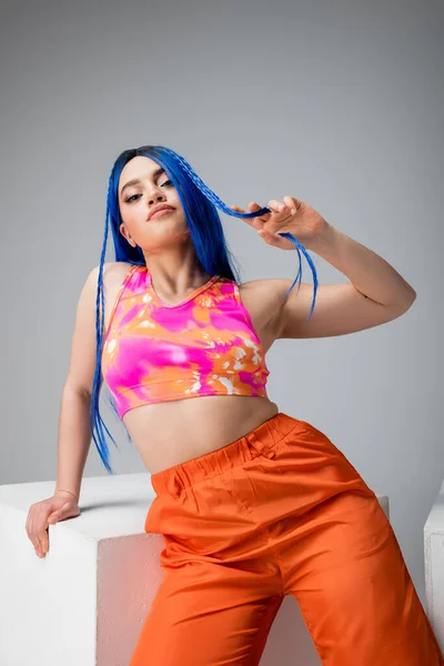 Style rebelle, jeune femme tatouée aux cheveux bleus posant en vêtements colorés près du cube blanc sur fond gris, regardant la caméra, individu moderne, mode urbaine, gen z — Photo de stock