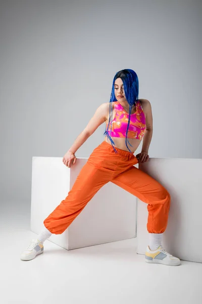 Стильный вид, татуированная молодая женщина с голубыми волосами позирует в красочной одежде рядом с белыми кубиками на сером фоне, полная длина, индивидуализм, современный стиль, городская мода, яркий цвет, модель — стоковое фото