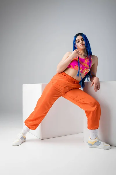 Modern subculture, tattooed woman with blue hair posing in colorful clothes near white cubes on grey background, full length, individualism, modern style, urban fashion, vibrant color, young model — Stock Photo