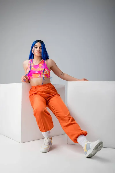Modern subculture, tattooed woman with blue hair posing in vibrant clothes near white cubes on grey background, full length, individualism, modern style, urban fashion, young model — Stock Photo