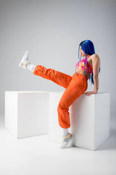 Tendências da moda, tatuado jovem mulher com cabelo azul posando com perna levantada perto de cubos brancos no fundo cinza, comprimento total, individualismo, estilo moderno, moda urbana, cor vibrante, modelo — Fotografia de Stock