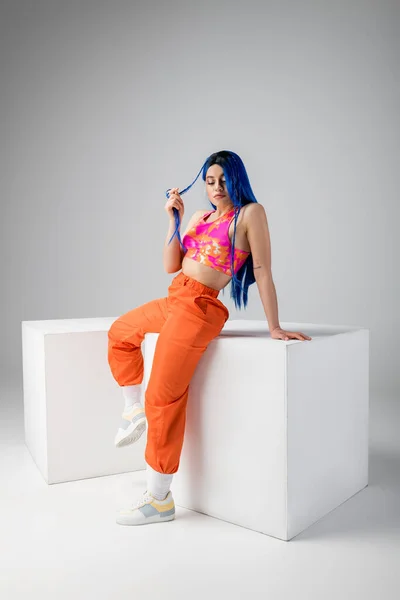 Fashion statement, tattooed young woman with blue hair posing in colorful clothes near white cubes on grey background, full length, individualism, modern style, vibrant color, urban fashion — Stock Photo