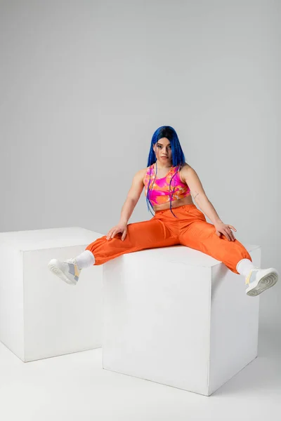 Fashion trends, tattooed young woman with blue hair sitting with outstretched legs on white cube on grey background, full length, individualism, modern style, urban fashion, vibrant color, model — Stock Photo