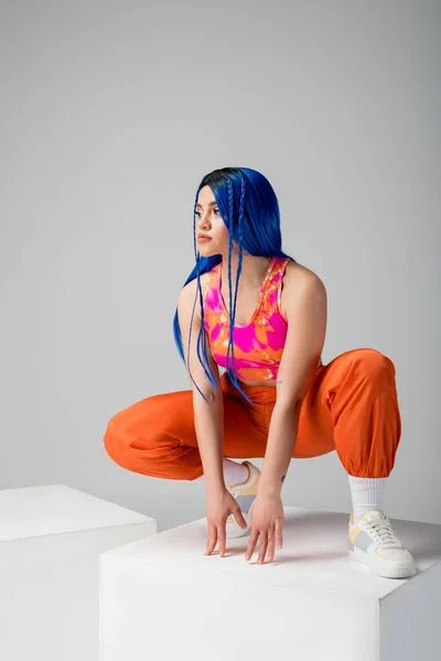 Moda e estilo, mulher jovem tatuada com cabelo azul sentado em cima do cubo branco no fundo cinza, comprimento total, individualismo, estilo moderno, moda urbana, cor vibrante, modelo — Fotografia de Stock