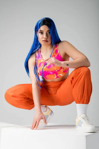 Moda juvenil, mujer joven tatuada con el pelo azul sentado en la parte superior del cubo blanco sobre fondo gris, longitud completa, individualismo, estilo moderno, moda urbana, color vibrante, modelo - foto de stock