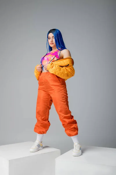 Full length, fashion forward, young woman with blue hair posing in colorful clothes on grey background, standing on white cubes, vibrant color, female model, individualism, urban fashion — Stock Photo